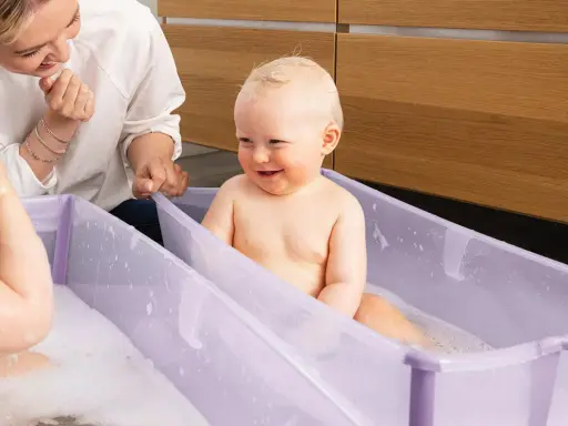 Glad mamma tittar på när ett litet barn njuter av att bada bebis i ett lila badkar med bubblor.