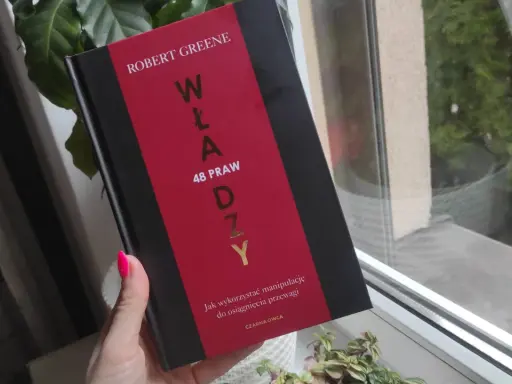 Dłoń z neonowym różowym lakierem trzyma książkę "48 praw władzy" Roberta Greene'a.