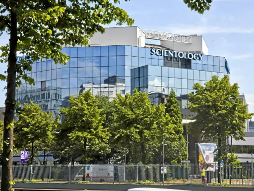 Bâtiment moderne aux façades vitrées reflétant le ciel bleu, avec le nom "SCIENTOLOGY" inscrit en lettres bleues lumineuses. Des arbres verdoyants entourent la structure.