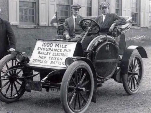 Un hombre junto a un coche eléctrico Bailey de 1907, parte de la historia del coche eléctrico, con un cartel que anuncia una carrera de resistencia de 1000 millas.