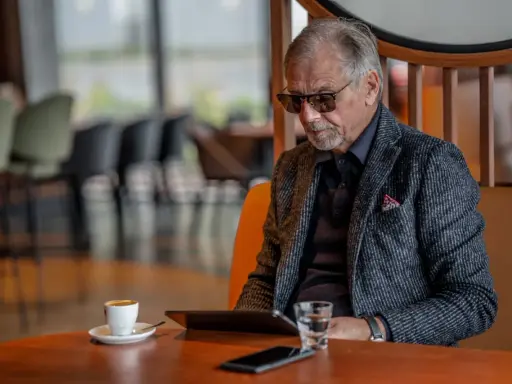 Elegancki starszy pan w okularach, z kawą i tabletem, zastanawia się, co kupić starszej osobie.