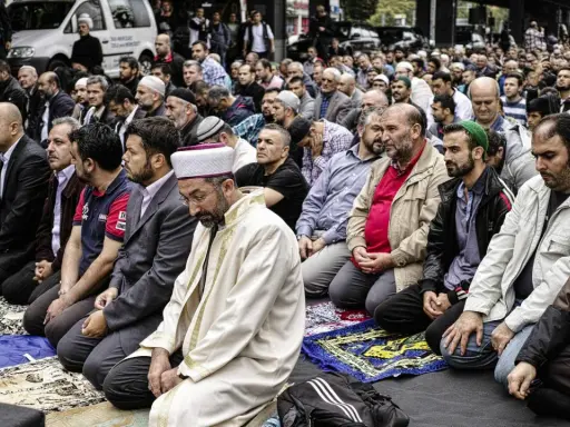 Männer in traditioneller Kleidung knien auf Gebetsteppichen. Ein Mann in einem weißen Gewand und Turban leitet das Gebet, ein Zeichen der berberischen Religion.