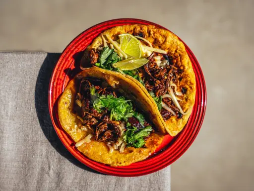 Zwei saftige Birria Tacos auf einem roten Teller, garniert mit Koriander und Limettenspalten.