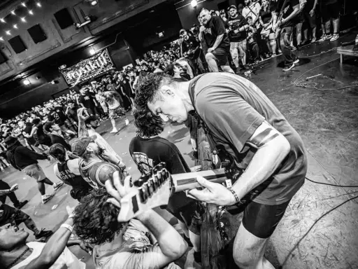 Energetyczny koncert w klubie. Czarno białe zdj. z tłumem szalejącym pod sceną, gdzie gitarzysta pochyla się nad instrumentem.
