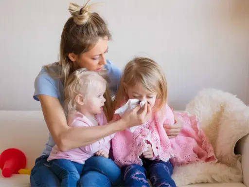 Mama wyciera nos chorej córce, podczas gdy młodsza siostra patrzy. Czy szczepienie mogłoby zapobiec katarowi?