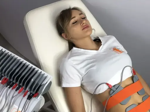 Kobieta leży na kozetce, poddana zabiegowi elektrostymulacji brzucha. Elektrody przymocowane są do jej talii.