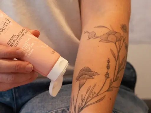 Une main tient un tube de crème réparatrice post-tatouage, prêt à être appliqué sur un bras orné d'un tatouage floral délicat.