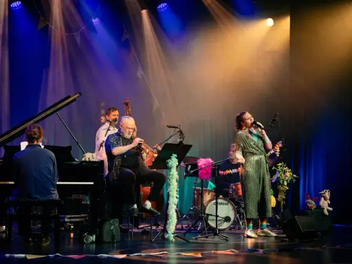En jazzgrupp uppträder på scenen under Stockholm Jazz Festival. En sångerska i glittrig klänning sjunger, en man spelar trumpet, en annan står vid basen och en person sitter vid pianot.