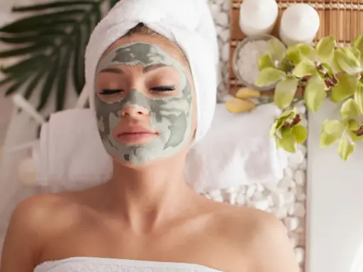 Femme détendue avec un masque fait maison, enveloppée dans une serviette, entourée de fleurs d'orchidée et de bougies.