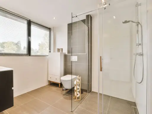 Salle de bain moderne avec une douche italienne, un WC suspendu et un meuble vasque. La lumière naturelle filtre à travers les stores.