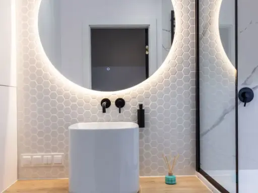 Baño moderno con lavabo blanco, espejo redondo con luz LED y azulejos hexagonales. ¿Cuánto cuesta reformar un baño así?