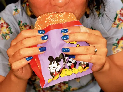 Mani con smalto blu intenso tengono un hamburger in un involucro con Topolino, in uno stile tipico di Martin Parr.