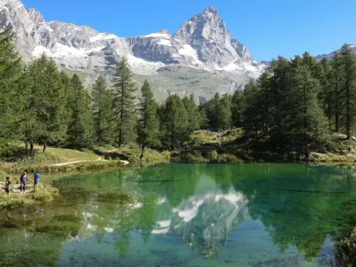 Un gruppo di escursionisti ammira il riflesso del Cervino nel lago blu di Cervinia, circondato da pini e montagne innevate.
