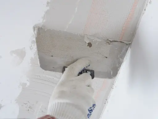 Ręka w rękawicy nakłada zaprawę na styropian przy suficie. Proces pokazujący, jak przykleić styropian do sufitu.