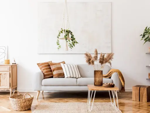 Un salón moderno con un sofá gris, cojines étnicos y una manta mostaza. La decoración se completa con una planta colgante y un jarrón con hierbas secas.