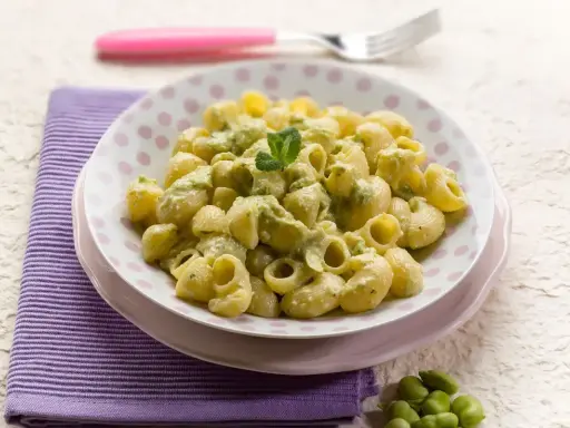 Piatto di pasta con le fave, condita con una crema verde e guarnita con menta. Un contorno di fave fresche completa la scena.