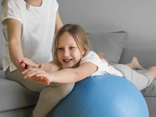 Dziecko z obniżonym napięciem mięśniowym ćwiczy mowę, leżąc na piłce rehabilitacyjnej pod opieką terapeuty.