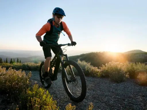 Kolarz w kasku i okularach jedzie rowerem po górskiej ścieżce. Słońce zachodzi za wzgórzami, tworząc piękne światło. To idealny moment na maraton MTB.