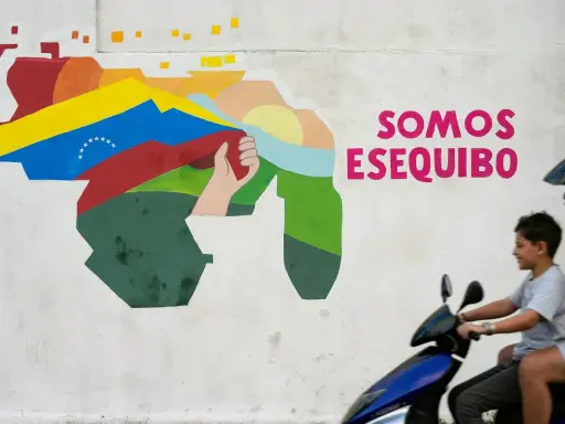 Mural przedstawiający mapę Wenezueli z flagą i hasłem "Somos Esequibo". Dwóch chłopców na skuterze przejeżdża obok.