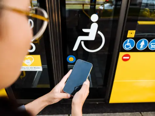 Osoba trzyma telefon przed drzwiami autobusu z symbolem wózka inwalidzkiego. To ułatwia podróżowanie, gdy masz legitymację osoby niepełnosprawnej.