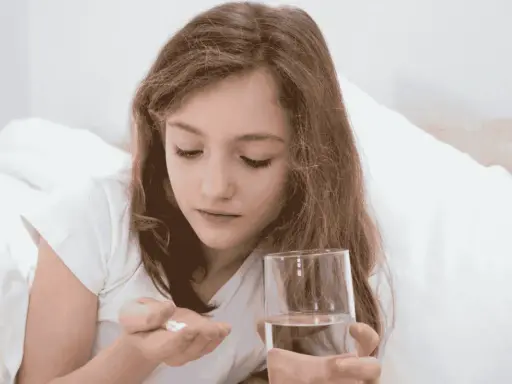 Dziewczynka w łóżku trzyma w dłoni tabletki i szklankę wody. Uważnie patrzy na białe tabletki, które mogą być przeterminowany paracetamol dla dzieci.