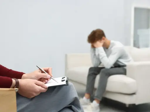 Młody mężczyzna siedzi na kanapie, ukrywając twarz w dłoniach, przeżywając cierpienie psychiczne. Obok niego terapeuta notuje coś na kartce.