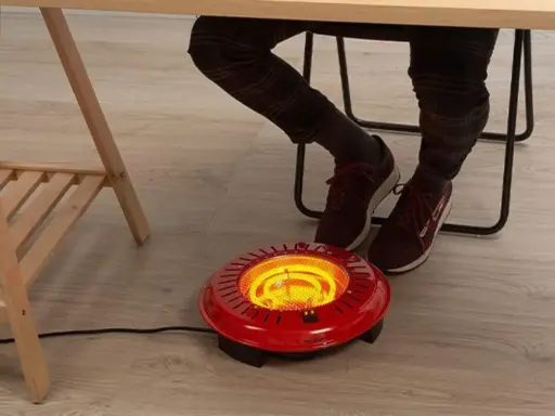 Persona calentando sus pies con un brasero eléctrico rojo bajo un escritorio.