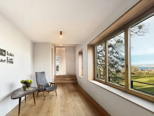 Interior moderno con suelo de madera, sillón gris y gran ventanal. El marco de madera de la ventana, que parece recién restaurado, enmarca un paisaje exterior.