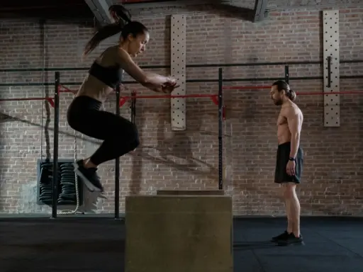 Frau springt auf eine Box, um ihre Kondition aufzubauen. Mann beobachtet sie im Fitnessstudio.
