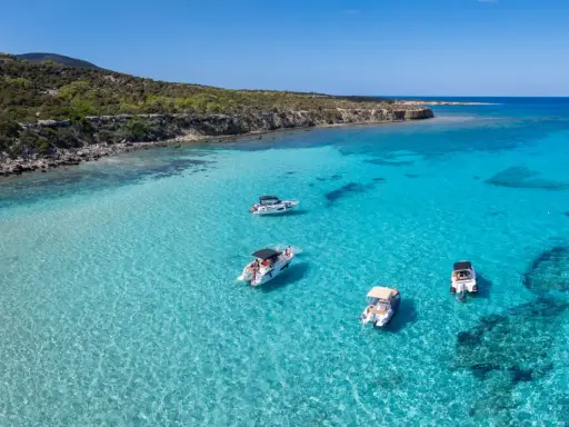 Traumhafter zypern strand mit kristallklarem Wasser und vier Booten, die vor der felsigen Küste ankern.