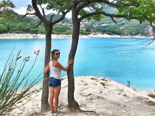 Donna sorridente abbraccia un albero con un lago turchese sullo sfondo. Una perfetta gita fuori porta nelle Marche.