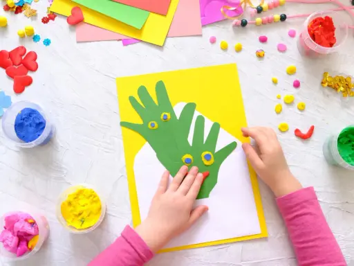 Kreatives Basteln mit Kleinkindern: Hände werden zu grünen Drachen auf gelbem Papier, umgeben von bunter Knete und Bastelmaterialien.