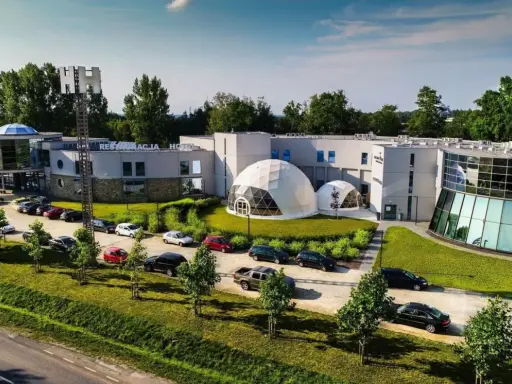 Nowoczesne polskie hotele niezależne z kopułami i przeszklonymi fasadami, otoczone zielenią i parkingiem pełnym samochodów.