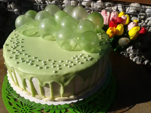 Zielony tort udekorowany koralikami i przezroczystymi, zielonymi balonami z żelatyny. Obok bukiet kolorowych tulipanów.