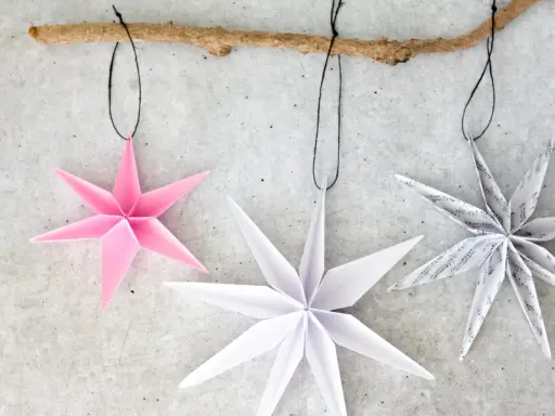 Drei Papiersterne, die Kinder falten könnten, hängen an einem Ast. Einer ist rosa, die anderen weiß und mit Noten bedruckt.