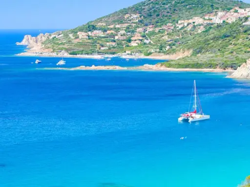 Un catamaran blanc avec des accents rouges navigue sur des eaux bleues cristallines, près d'une côte verdoyante et d'un village pittoresque.