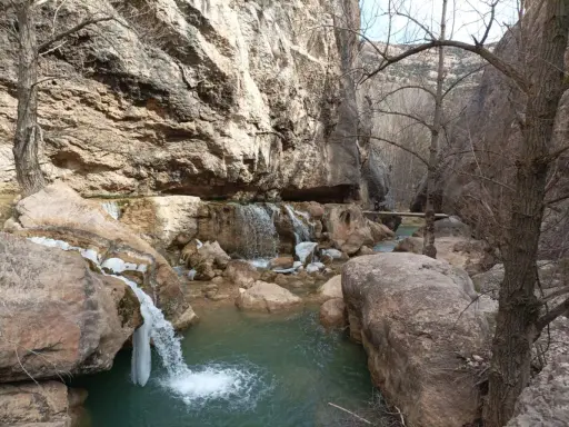 Cascada y río en un cañón rocoso, con hielo en las rocas. Un puente de madera cruza el río, evocando las pasarelas de Valloré.