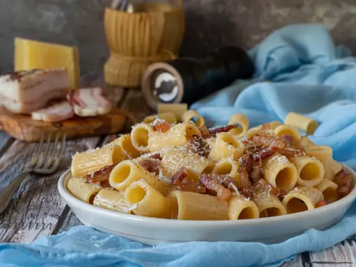 Piatto di pasta alla gricia fumante, guanciale croccante e pecorino romano grattugiato. Ingredienti freschi su un tavolo rustico.