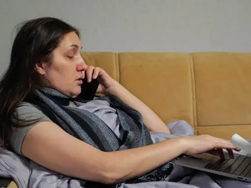 Kobieta w łóżku, rozmawiająca przez telefon i pracująca na laptopie, zastanawia się, na ile dni opłaca się brać zwolnienie lekarskie.