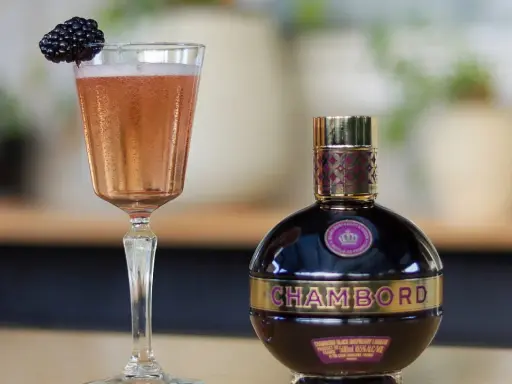 Elegancki kieliszek z różowym drinkiem i jeżyną obok butelki likieru Chambord.