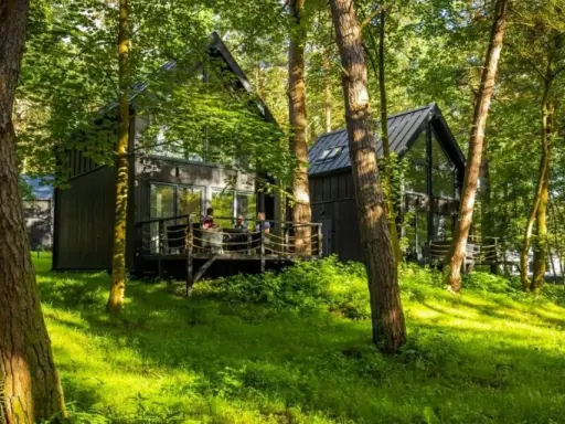 Nowoczesne **hotel w lesie**, z drewnianymi tarasami i dużymi oknami, otoczony zielenią.