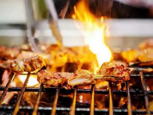 Marinierte Lammkoteletts brutzeln auf dem Grill. Flammen züngeln über dem Fleisch, das perfekt gegrillt wird.