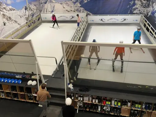 Ludzie ćwiczą jazdę na nartach na sztucznym stoku narciarskim w Ski Arenie Wrocław. Widać sprzęt narciarski i buty narciarskie w schowkach.