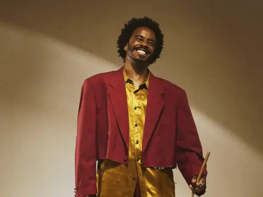 Un percussionniste célèbre, souriant, porte une veste courte bordeaux et une chemise dorée, tenant une baguette.