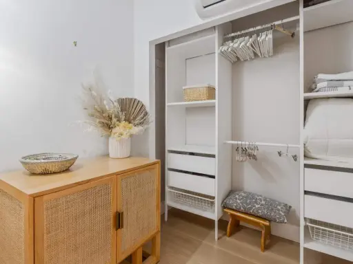 Un armario blanco organizado, listo para limpiar armarios por dentro. Cestas, ropa de cama y un jarrón con flores secas decoran el espacio.