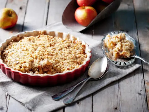 Un délicieux crumble pomme individuel, doré et croustillant, prêt à être dégusté. Des pommes fraîches sont visibles en arrière-plan.