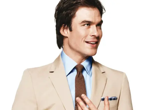 A man in a tan suit and blue shirt smiles, adjusting his tie.