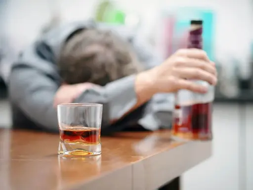 Zaniedbany mężczyzna, prawdopodobnie po eksmisji alkoholika z domu, leży na stole obok szklanki z whisky i butelki.