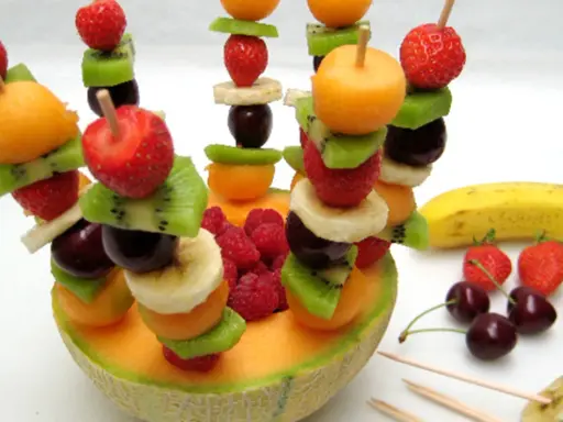 Une **brochette de fruits** colorée avec fraises, kiwis, bananes, cerises et melon, présentée dans une moitié de melon.