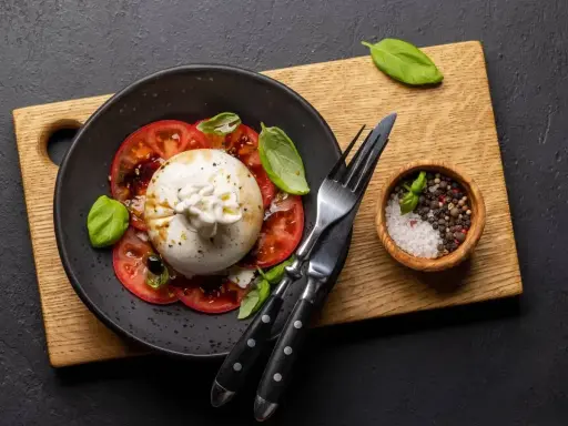Pyszna burrata z pomidorami, polana oliwą, udekorowana bazylią. Obok miseczka z solą i pieprzem.